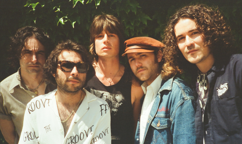 Five men with long hair stand close together outdoors in front of green foliage, looking at the camera. 