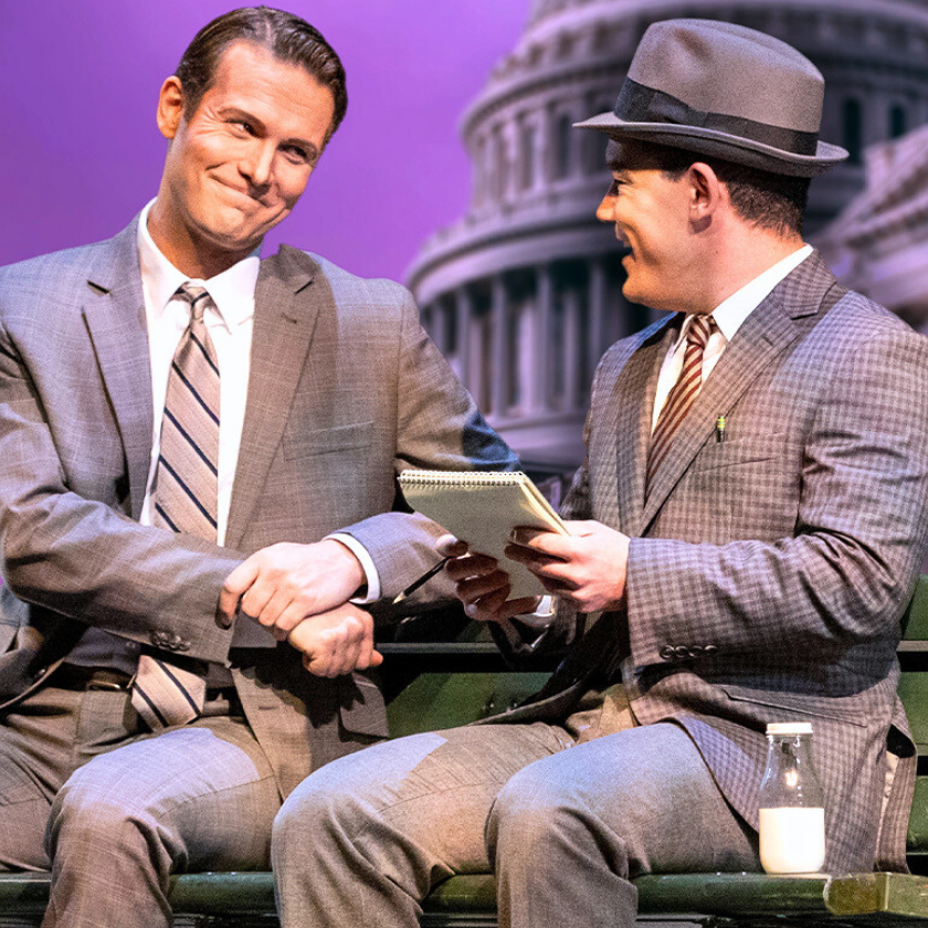 Two men in suits sit on a green bench, smiling and talking in front of a backdrop featuring the U.S. Capitol. One holds a notebook and pen, the other gestures with his fist; a milk bottle sits beside them.
