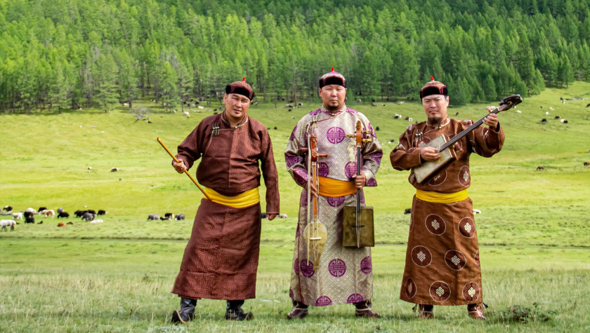 Three men in traditional Mongolian clothing stand on a grassy field with trees and grazing animals in the background. One holds a stringed instrument, and all wear hats and bright yellow sashes.