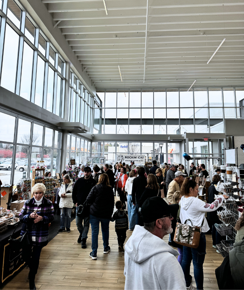 A large group of people shop at an indoor market filled with vendor tables under high ceilings and tall windows, with natural light streaming in. 