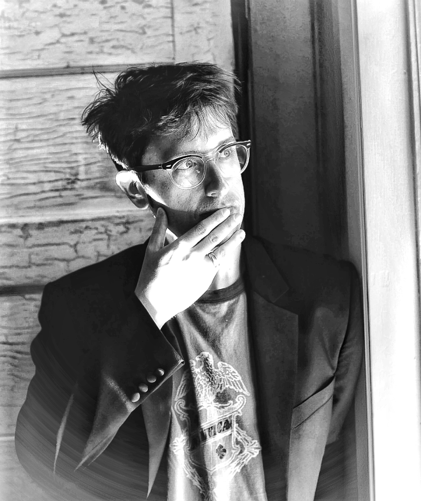 A person with glasses stands by a window, touching their mouth thoughtfully.