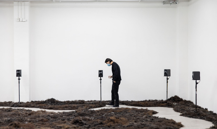Person wearing a mask stands in a white room with speakers and dark material on floor.