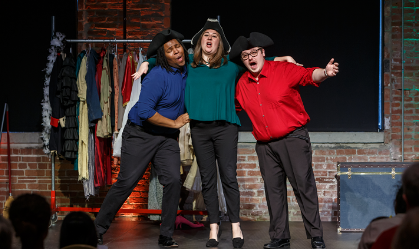 Three people on stage in costume hats sing with arms around each other.