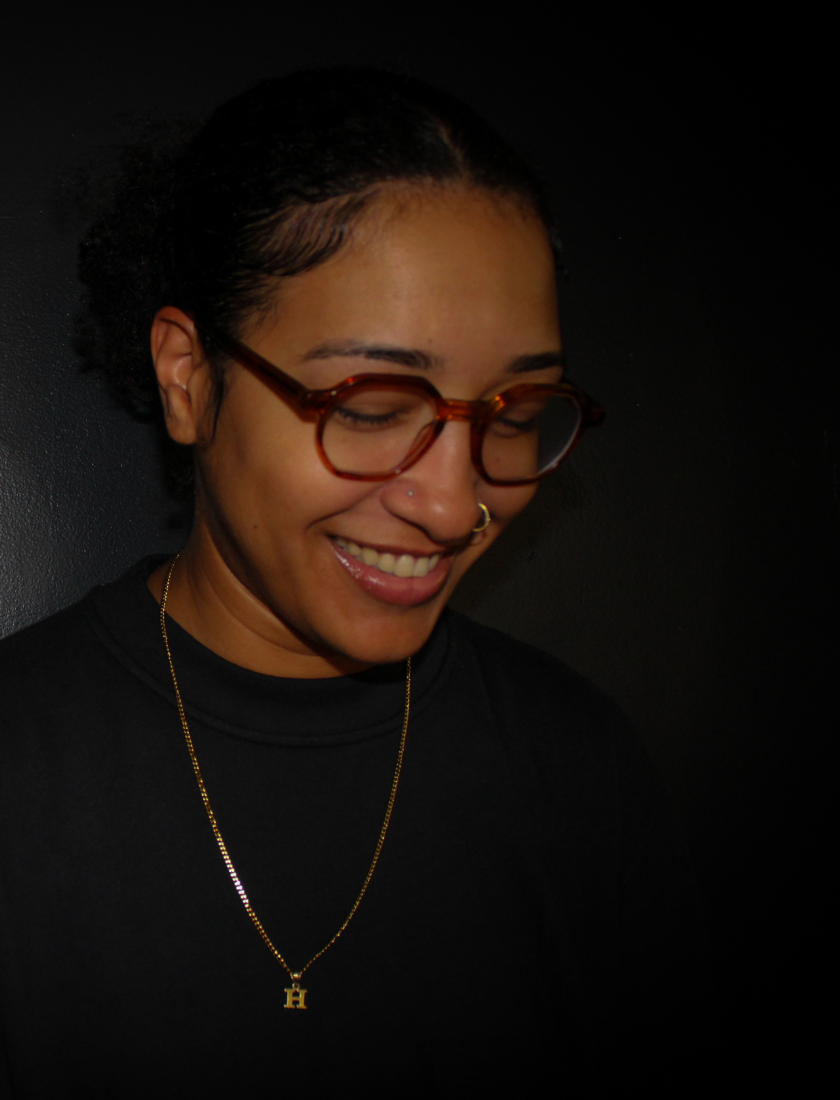Person with glasses and necklace smiling, looking down against a dark background.