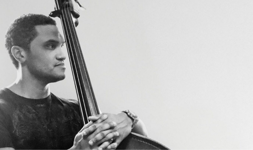 A man with short hair looks to the left while holding an upright bass, resting his hands on top of the instrument, against a plain light background.