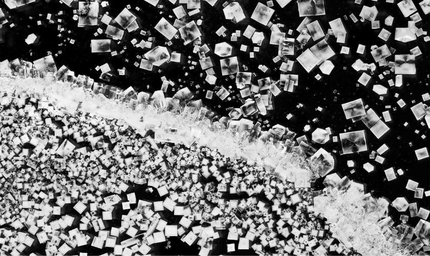 Close-up image of salt crystals scattered on a dark surface, forming a dense cluster along a diagonal line and more dispersed individual crystals above it.