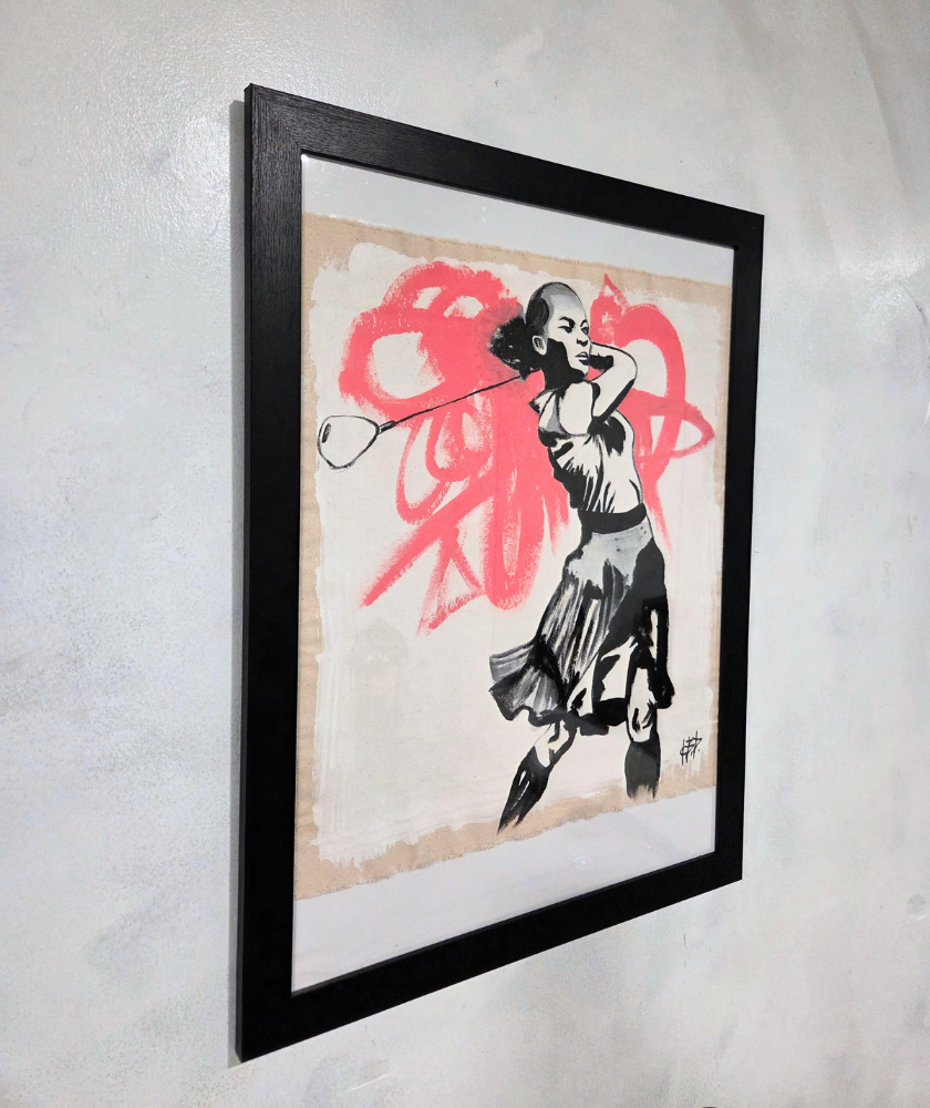Framed artwork of a girl swinging a golf club, painted in black and white with a bold red abstract swirl in the background, hanging on a light-colored wall.