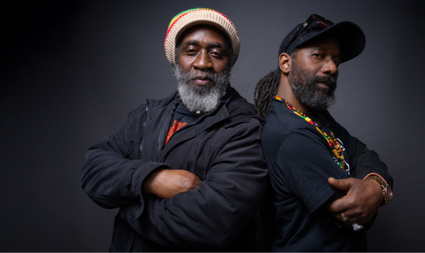 Two men stand back to back with arms crossed, both wearing black shirts. One wears a knit hat and jacket, the other a cap and beaded necklaces. They both have beards and confident expressions against a dark background.