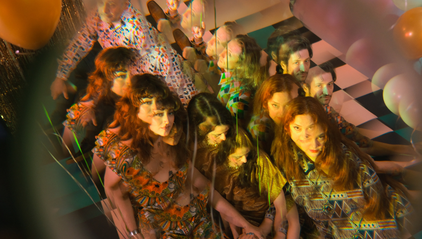 A group of people in colorful patterned outfits gather close together, with a prismatic lens effect creating multiple overlapping images of their faces and bodies. Balloons and checkered flooring are visible in the background.