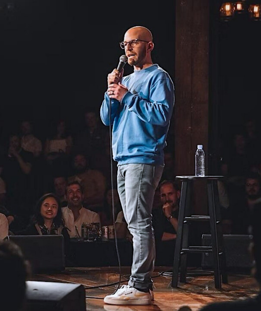A man wearing glasses, a blue sweatshirt, and jeans stands on stage holding a microphone. He is performing stand-up comedy in front of a seated audience, with a stool and water bottle beside him.