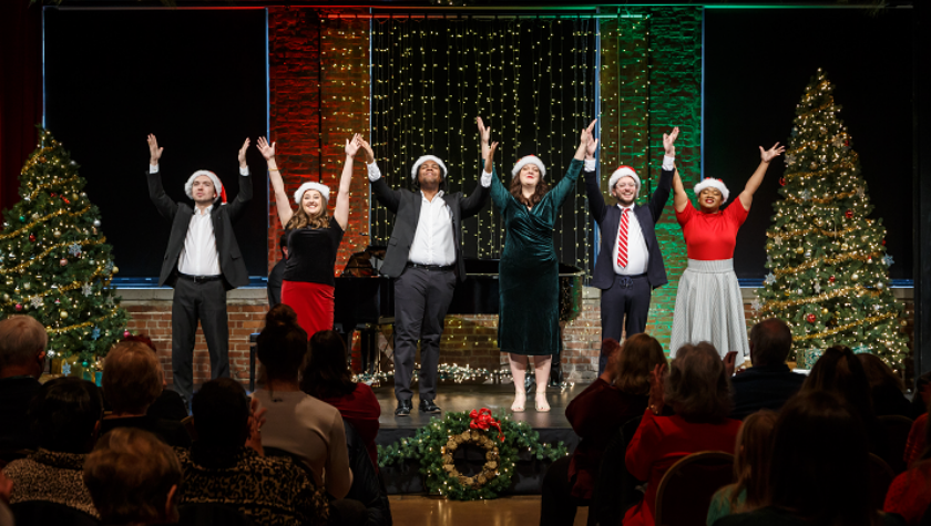 Six people in Santa hats perform on stage, arms raised, in front of an audience.