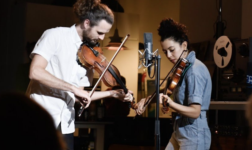 Two people playing violins in front of a microphone in a dimly lit room.