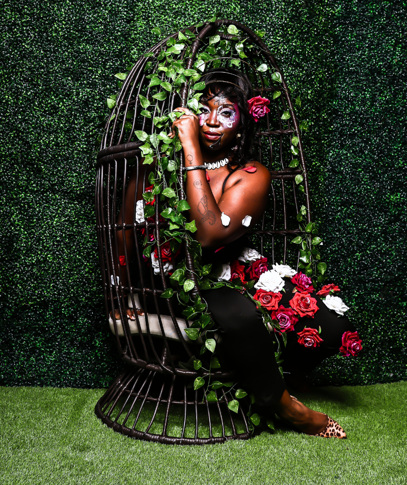 Person poses in a leafy hanging chair adorned with red and white roses, green background.