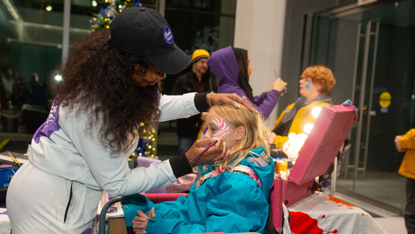 Woman paints a childs face at a festive indoor event with others in the background.