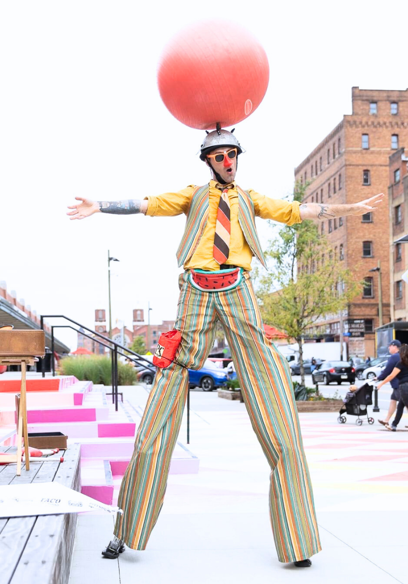 A performer on stilts balances a red ball on a helmet in an urban area.