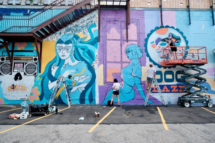 Four artists work on a colorful mural featuring stylized women and bold designs on a large exterior wall, using ladders and lifts. Paint cans and supplies are scattered in the parking lot below.