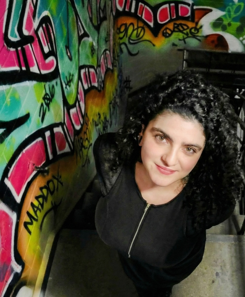 Woman with curly hair stands by graffiti-covered wall, looking up at the camera.