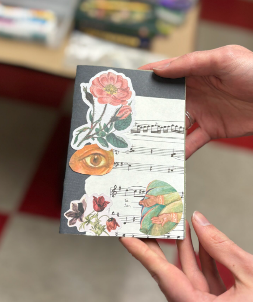 A hand holds a collaged card with flowers, an eye, and sheet music.
