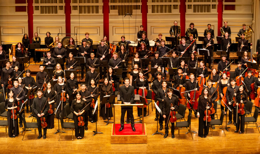 A full orchestra poses on stage with their conductor in a concert hall.