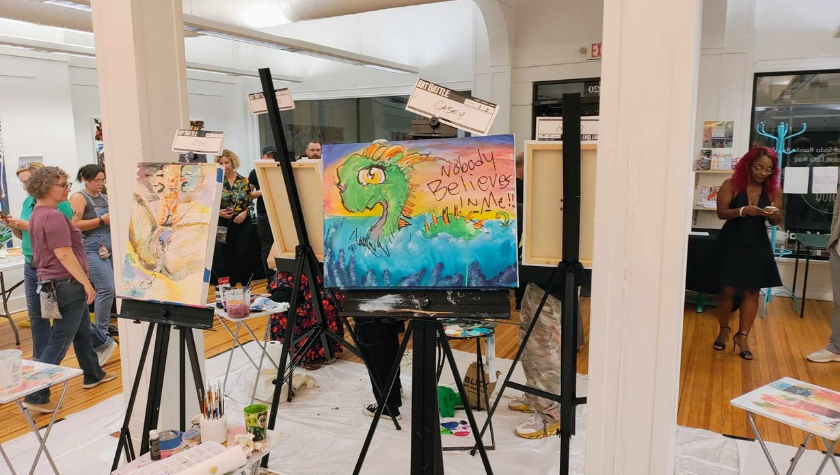 Several people paint on canvases in a well-lit art studio with easels and supplies.