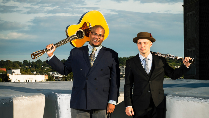 Two men in suits stand outside; one holds a guitar, the other holds a clarinet.