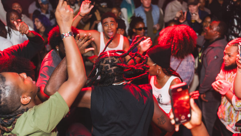 A crowd of people dance and raise their hands at a lively indoor party.