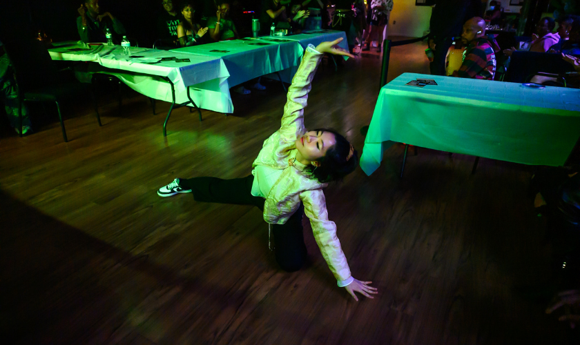 A person poses on a wooden floor in front of tables at an indoor event.