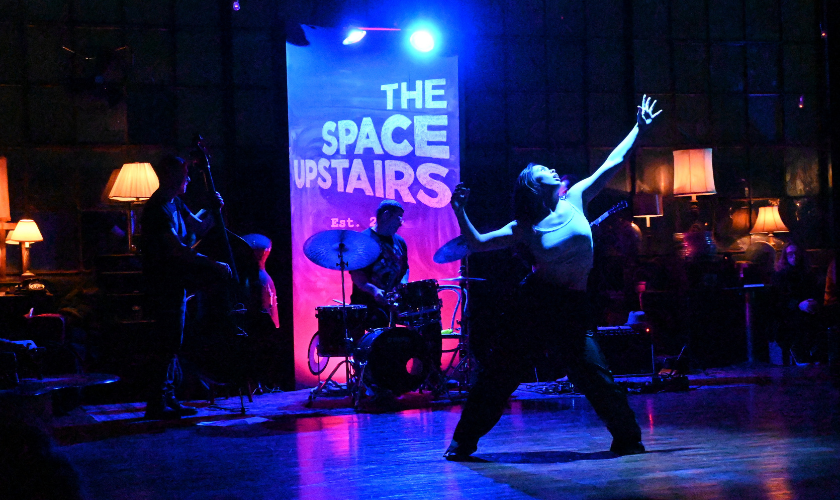 A dancer performs on stage with a band under blue and purple lighting.