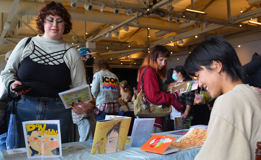 People browse and display comics at a table during an indoor comic convention.