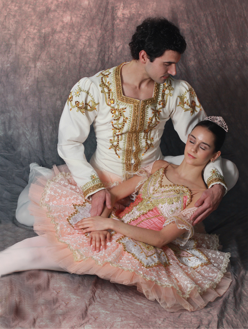 A male and female ballet dancer in ornate costumes pose in a seated embrace.