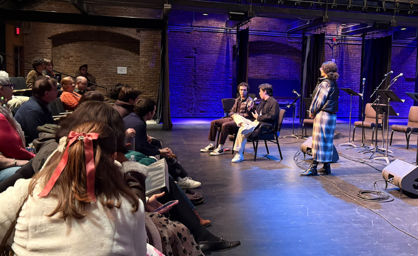 Three people speak on stage to an audience in a theater with blue lighting.