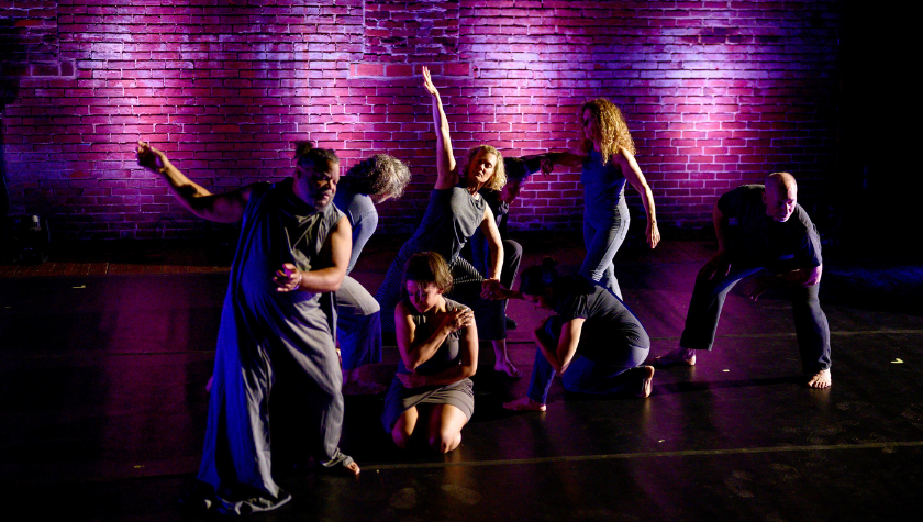 Seven people perform expressive dance poses on a stage with a lit brick wall.