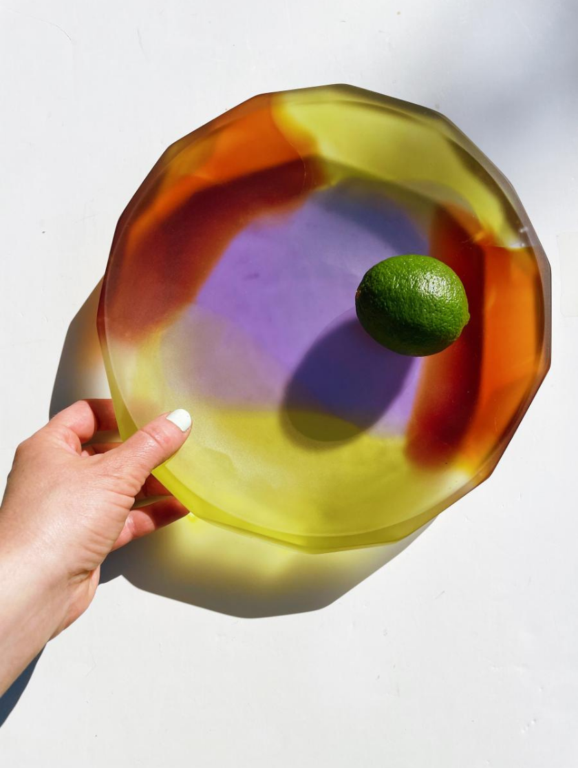 Hand holding a translucent, colorful plate with a lime placed inside on a white surface.