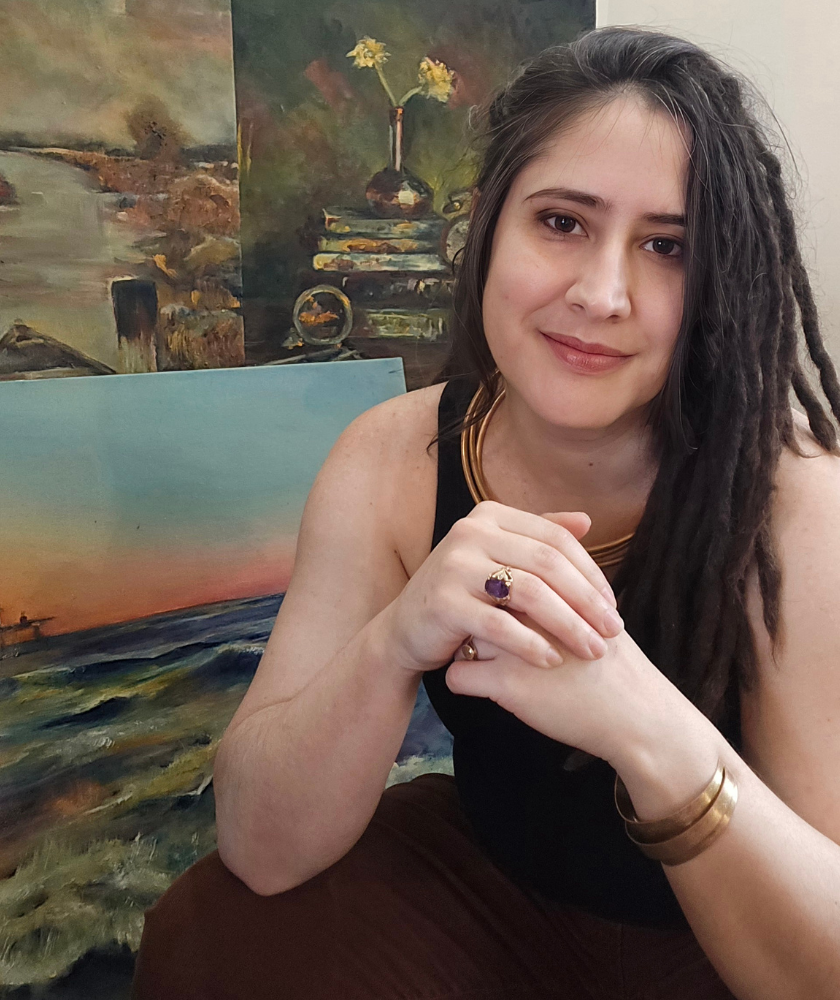 Woman with long hair sits in front of several landscape paintings, smiling.