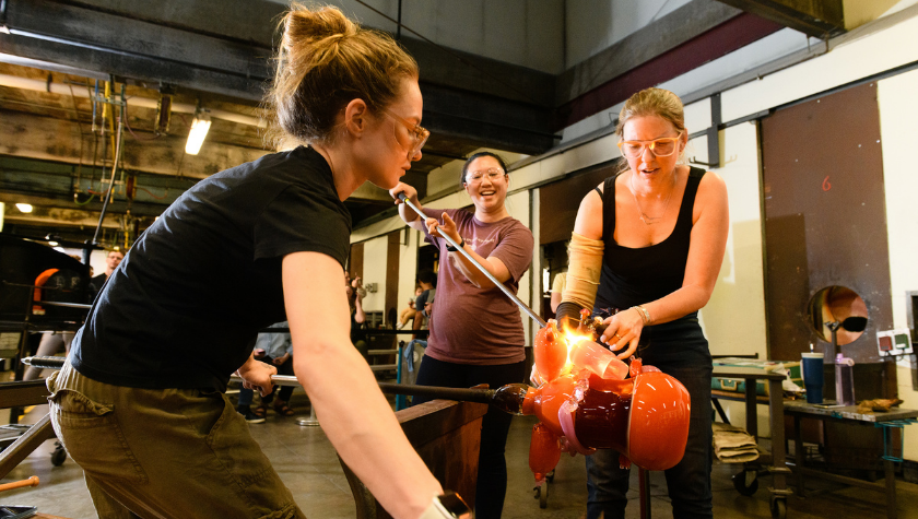 Three women work together shaping hot glass in a glassblowing studio.