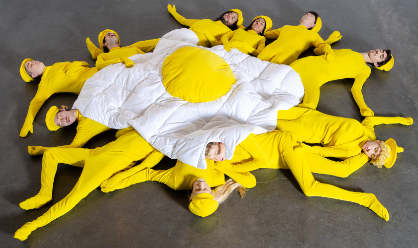 People in yellow bodysuits form the shape of a fried egg on a gray floor.