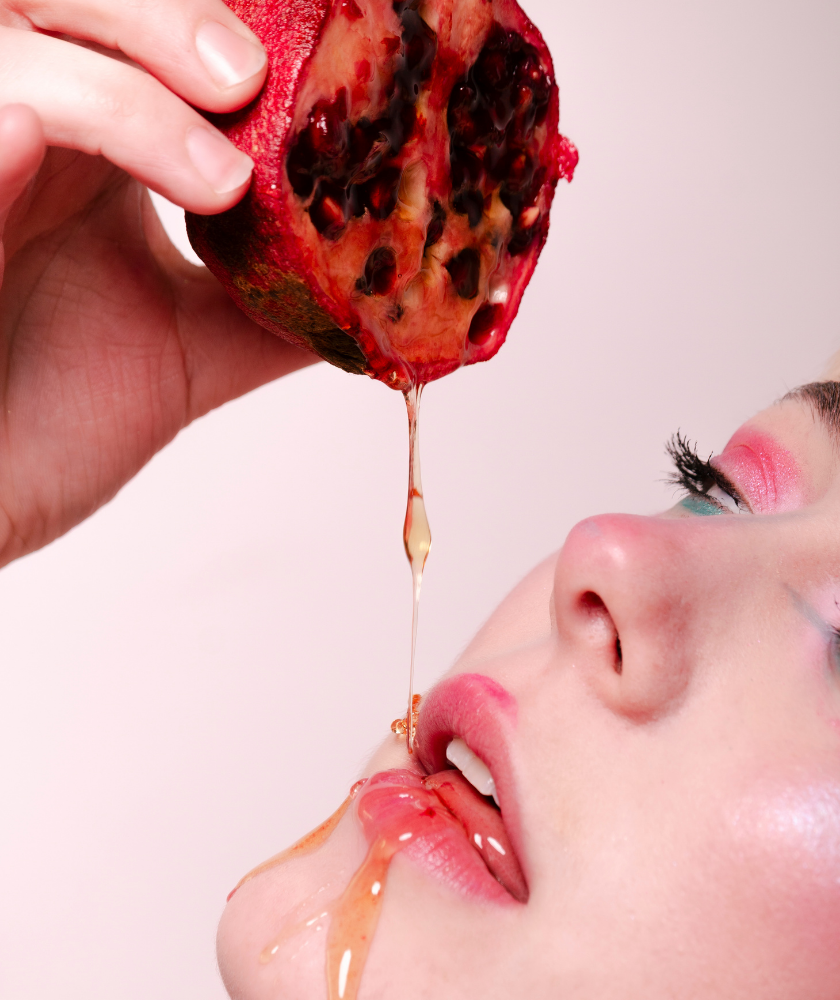 Juice drips from a pomegranate onto a womans face with colorful eye makeup.