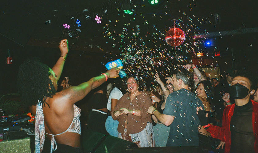 A lively crowd dances as a dancer sprays bubbles at a nightclub with colorful lights.