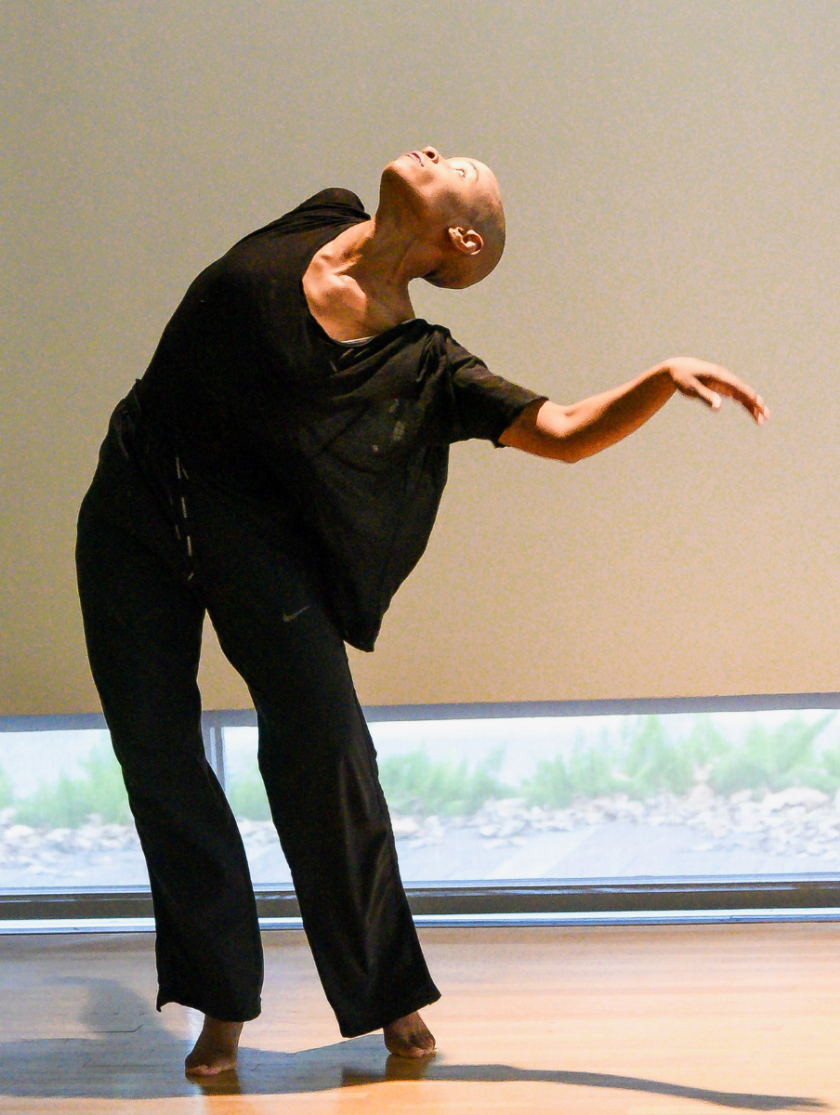 A dancer in black poses gracefully indoors with one arm extended and head tilted back.