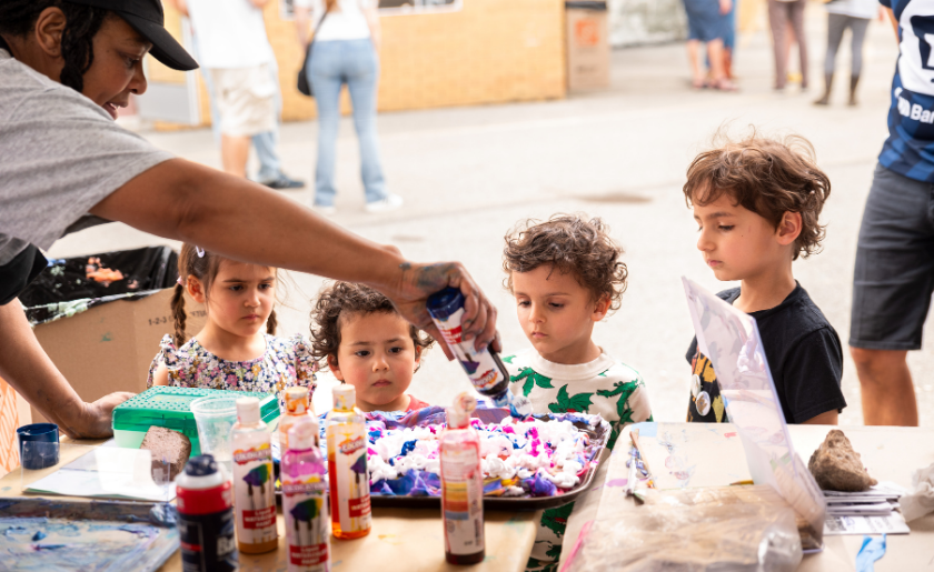 An adult helps four children with a colorful painting activity at a table outdoors.