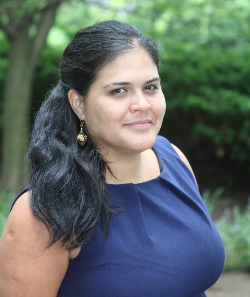 Woman with long dark hair in a blue top, standing outdoors and smiling slightly.