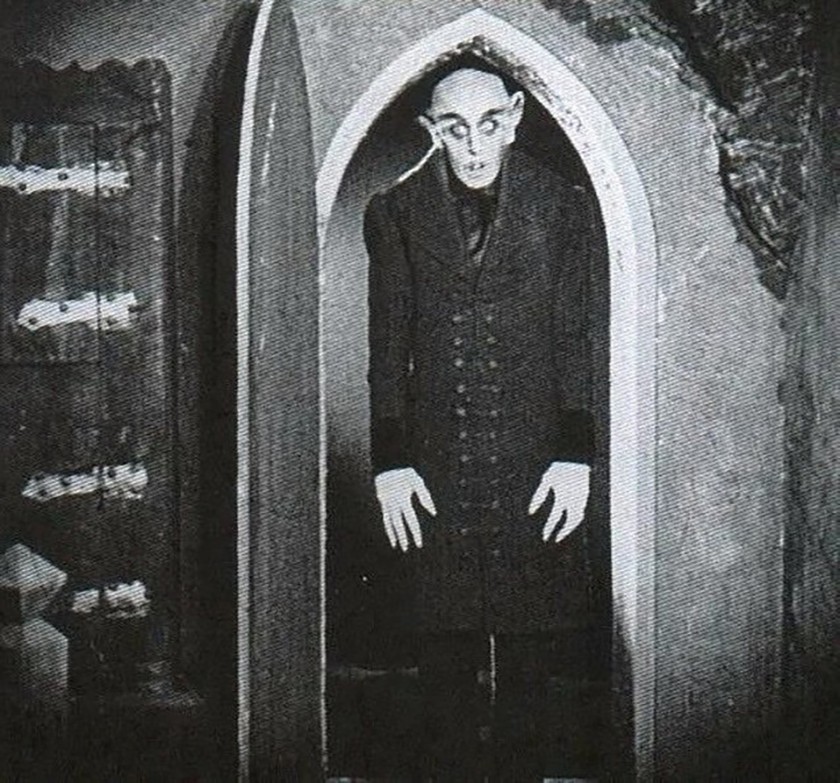 A pale, gaunt figure with pointed ears and long fingers stands in a dark arched doorway, dressed in a long, buttoned coat. The image is in black and white, evoking a classic horror film atmosphere.
