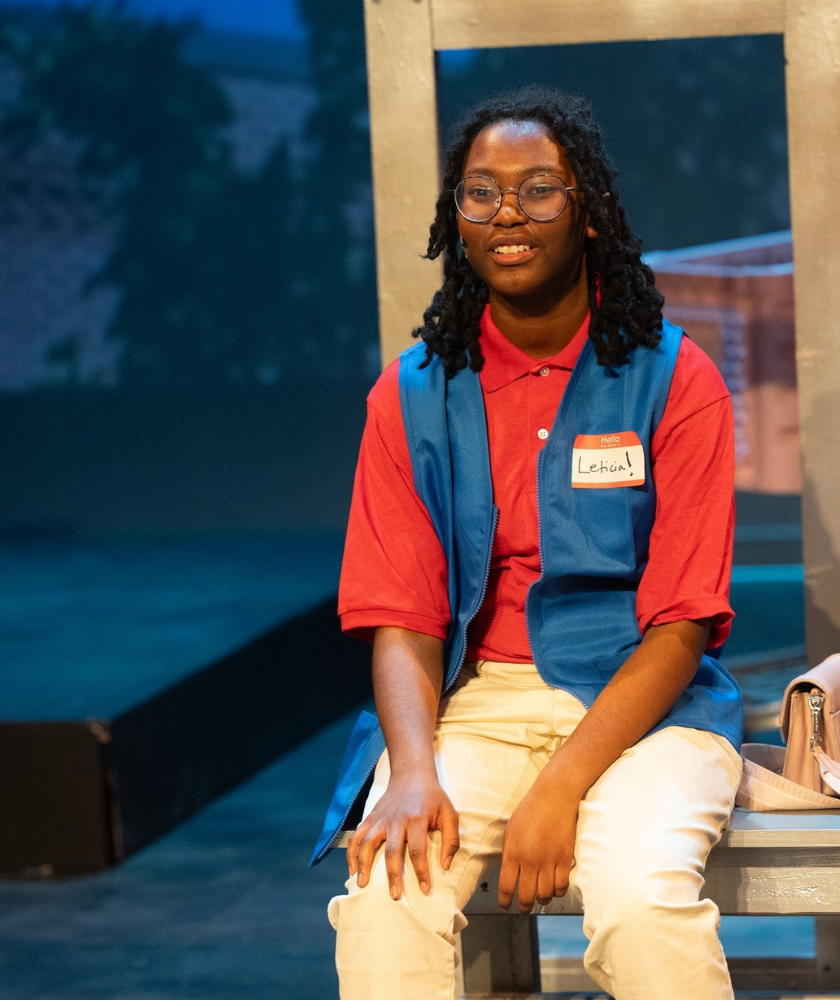 Person with glasses and name tag Lehia sits on a bench, wearing a red shirt and blue vest.