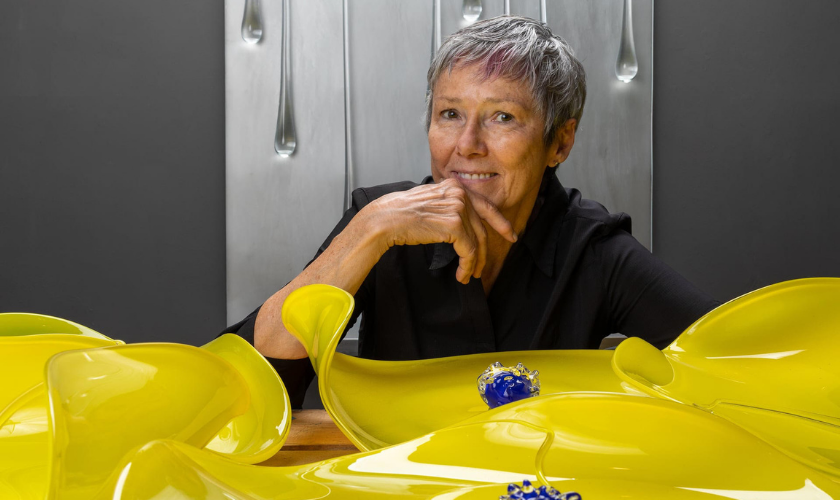 A person sits behind yellow glass art pieces, smiling with hand on chin.