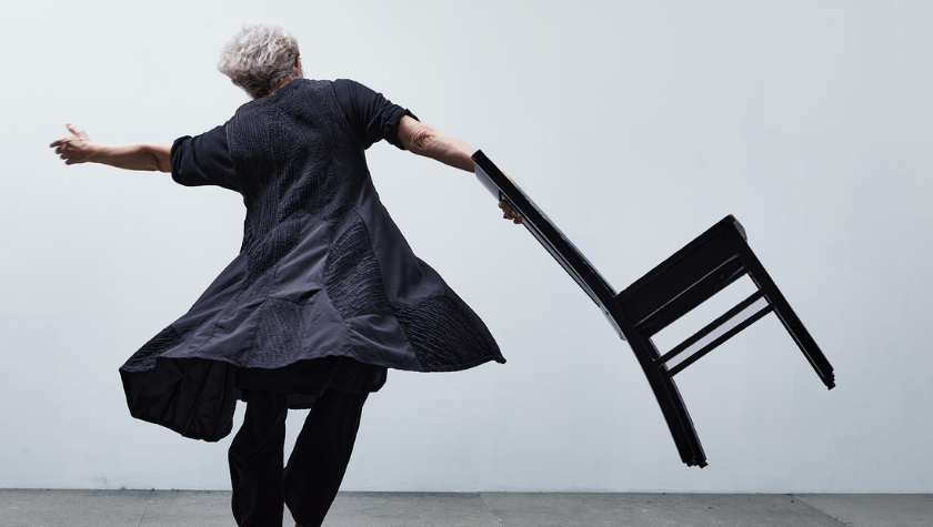 Person in black outfit spins while holding a chair against a plain, light background.