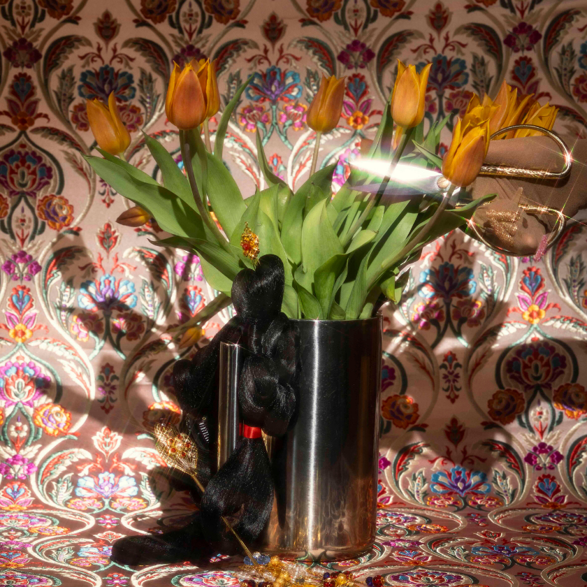 Yellow tulips in a metal vase with hair extensions, jewelry, and gloves on patterned background.