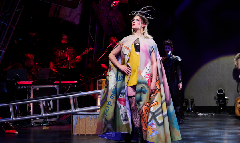 Person in colorful, graffiti-style cape poses on stage with musicians and dramatic lighting.