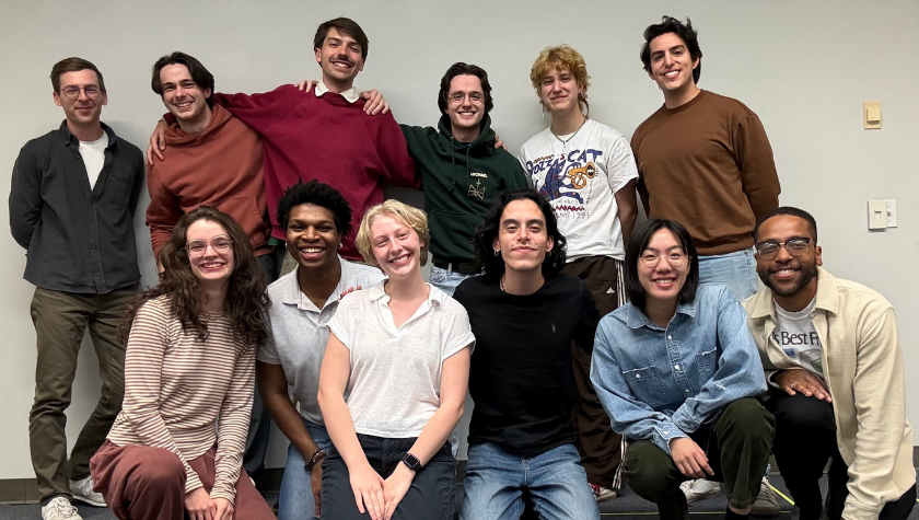 Twelve smiling people pose together in two rows in front of a plain wall.
