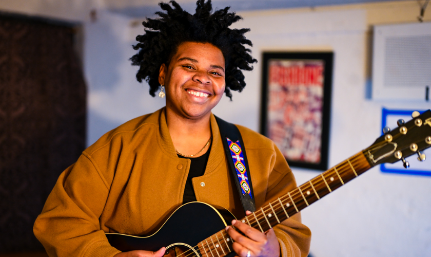 Person smiling and playing a black acoustic guitar indoors, wearing a brown jacket.