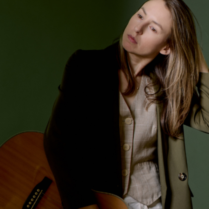 Person holding a guitar, tilting head, and touching hair, standing against a green background.